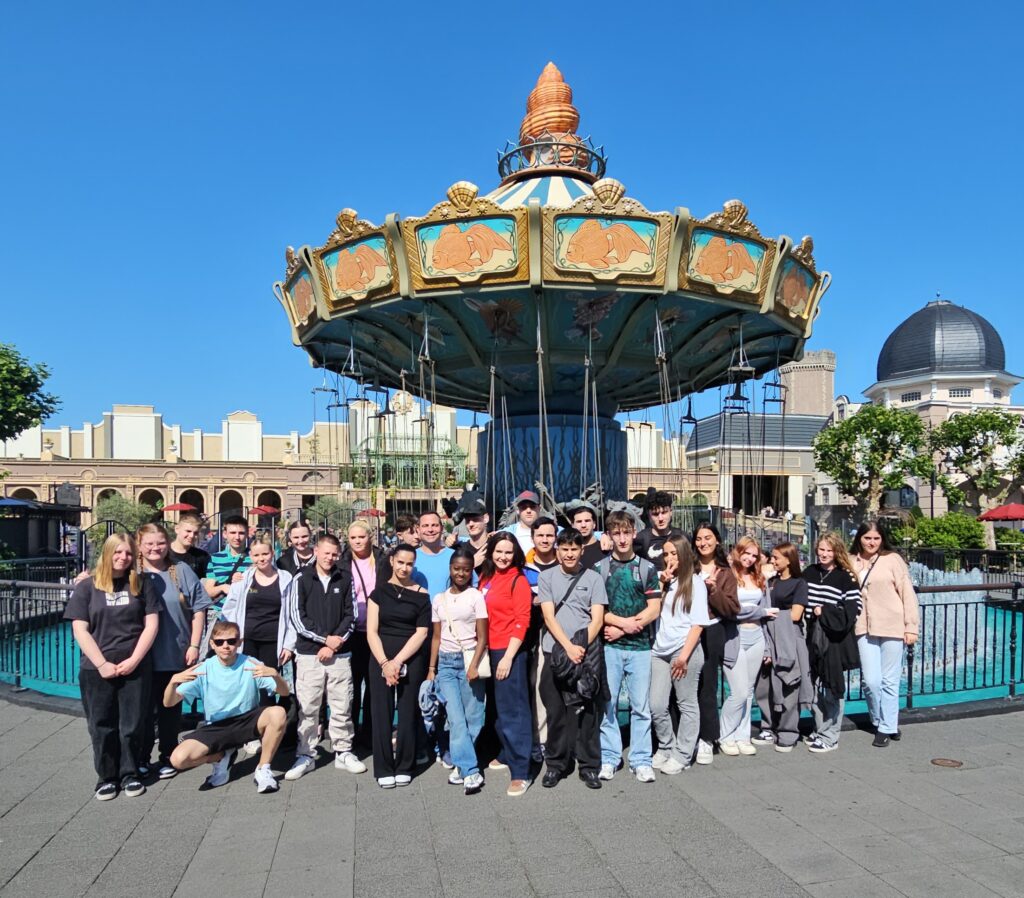 Gruppe von etwa 30 Jugendlichen steht vor einem Kettenkarussell in einem Freizeitpark, alle blicken in die Kamera.
