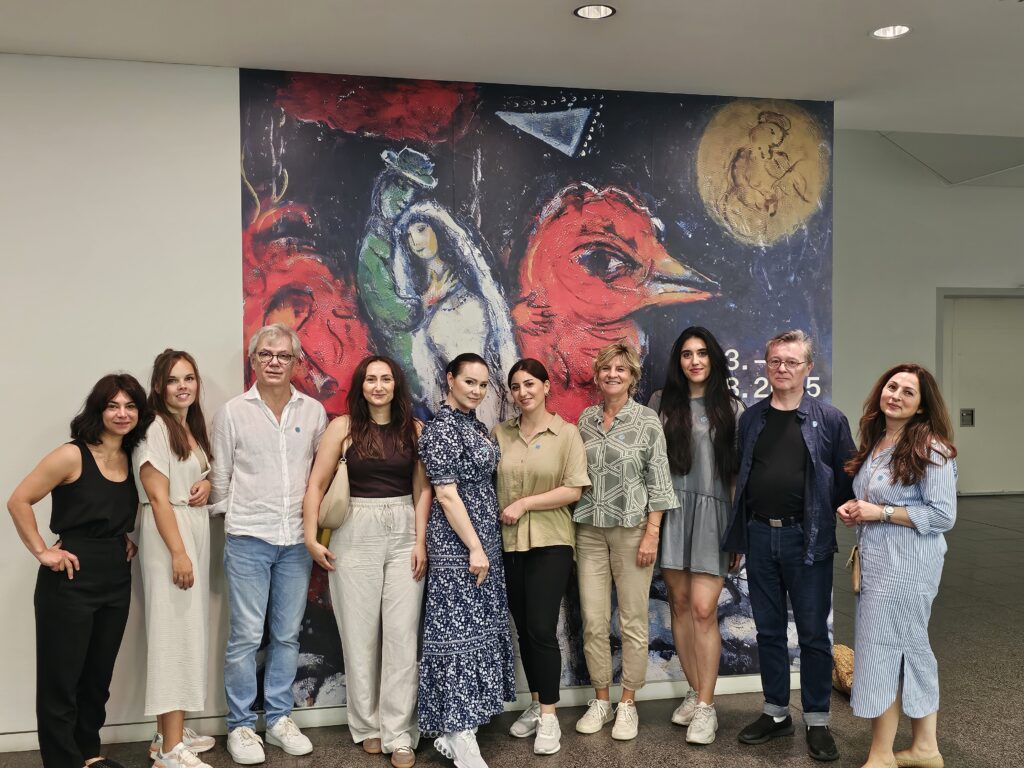 Gruppe von zehn Erwachsenen steht vor einem bunten Wandbild mit rotem Vogel und umarmendem Paar. Alle blicken in die Kamera.