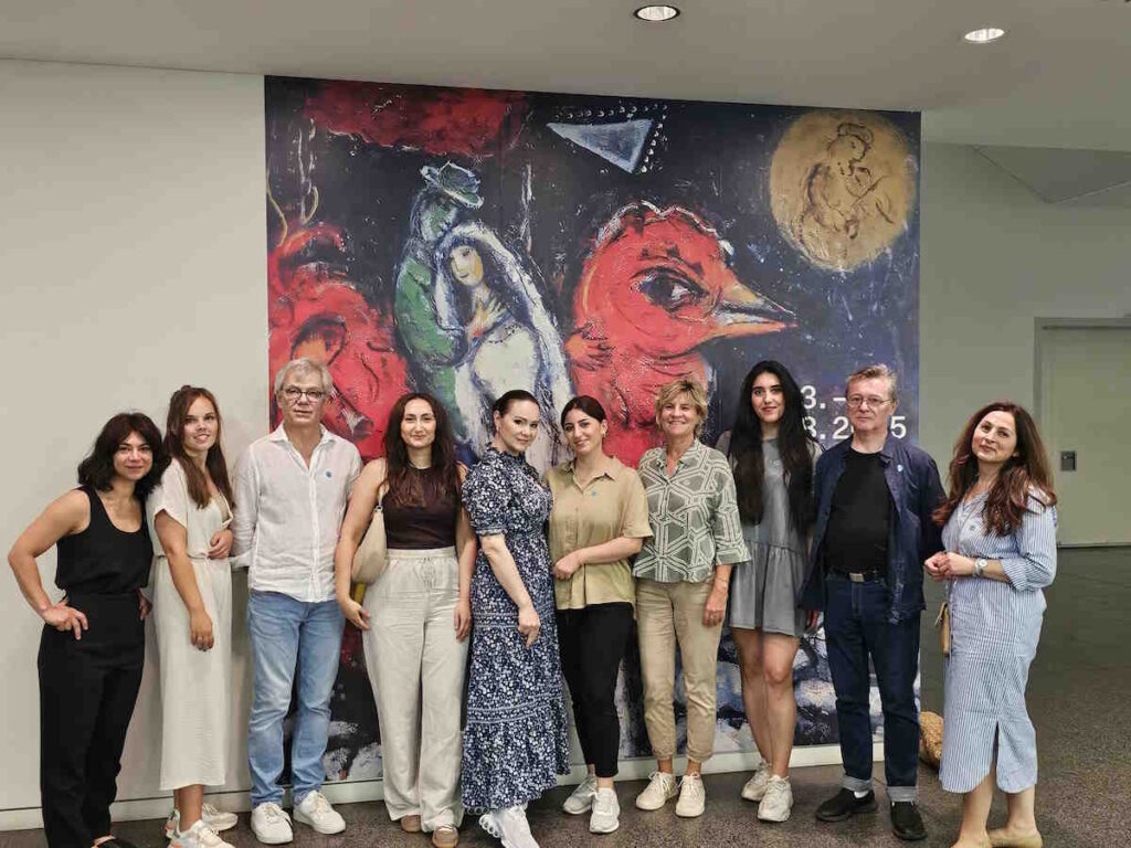 Gruppe von zehn Erwachsenen steht vor einem bunten Wandbild mit rotem Vogel und umarmendem Paar. Alle blicken in die Kamera.