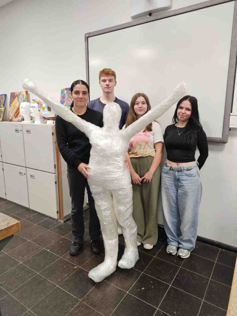 Vier Jugendliche stehen in einem Klassenraum neben einer großen menschlichen Figur, die aus transparenter Folie gestaltet wurde.