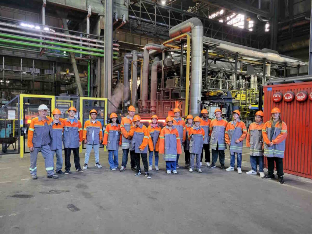 Gruppe von Jugendlichen und Erwachsenen in Schutzkleidung und Helmen steht in einer Industriehalle vor einer technischen Anlage.