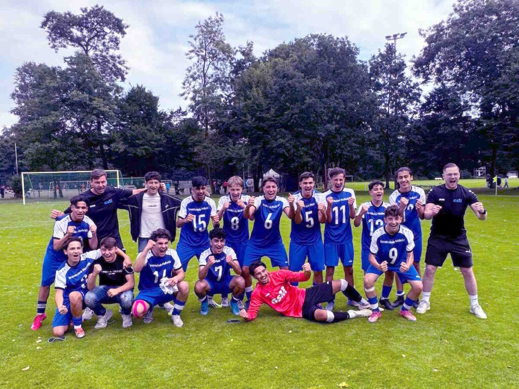 15 Schüler der U17-Fußballmannschaft der GSM jubeln nach dem gewonnenen Endspiel auf dem Rasenplatz gemeinsam mit ihren beiden betreuenden Sportlehrern.