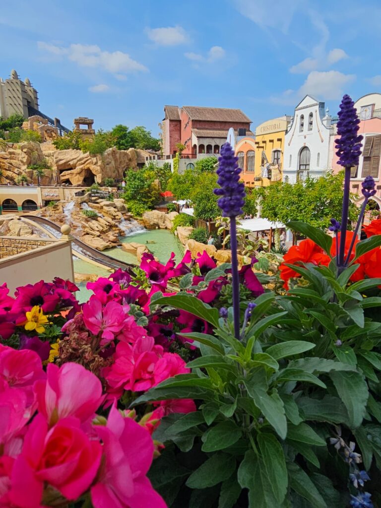 Bunte Blumen im Vordergrund, dahinter eine Themenwelt mit Wasserbahn, Felsen und Gebäuden im Stil eines Freizeitparks bei sonnigem Wetter.