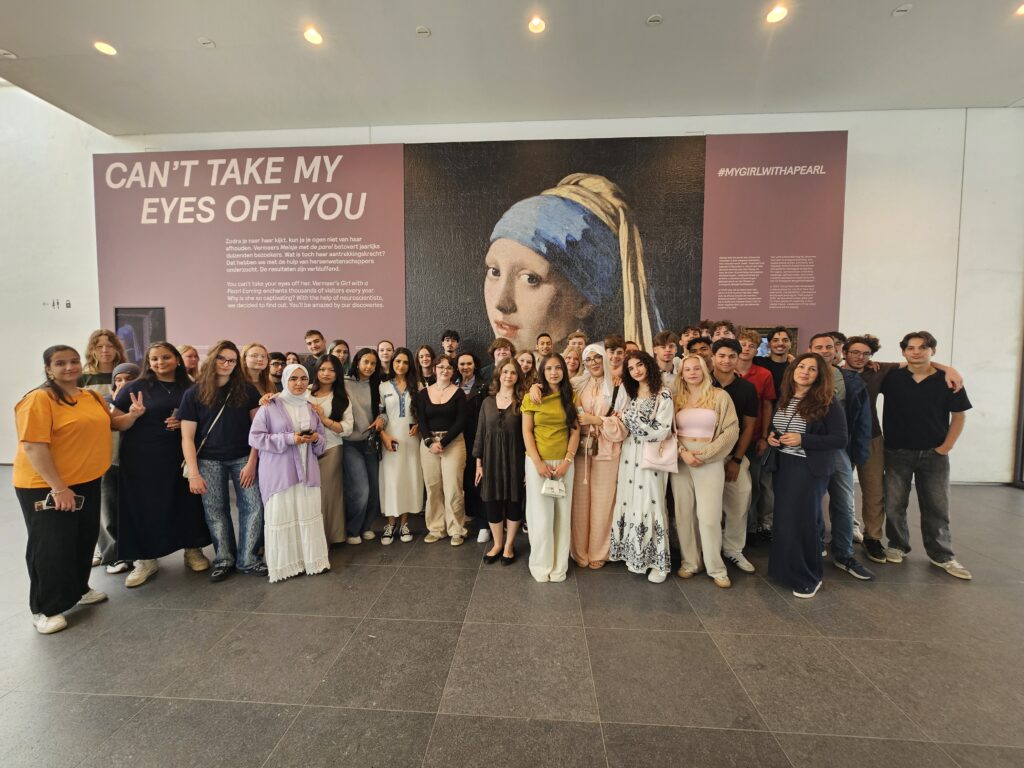 Eine große Gruppe Jugendlicher und Erwachsener steht vor einem Ausstellungsplakat mit dem Bild „Mädchen mit dem Perlenohrring“ und dem Text „CAN'T TAKE MY EYES OFF YOU“. Alle blicken in die Kamera.