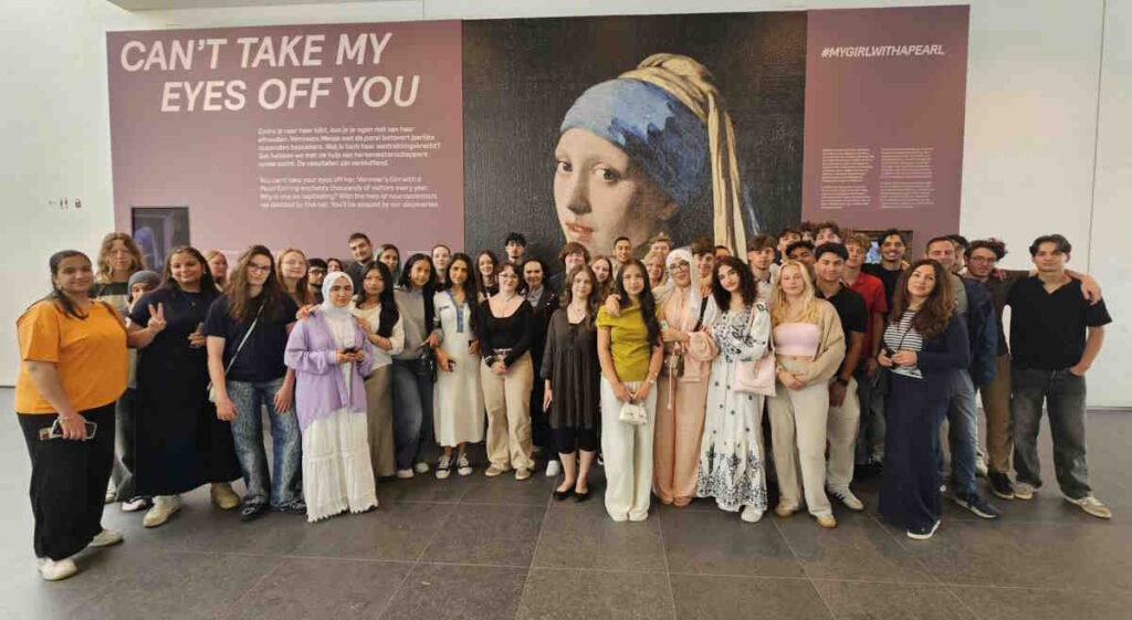 Eine große Gruppe Jugendlicher und Erwachsener steht vor einem Ausstellungsplakat mit dem Bild „Mädchen mit dem Perlenohrring“ und dem Text „CAN'T TAKE MY EYES OFF YOU“. Alle blicken in die Kamera.