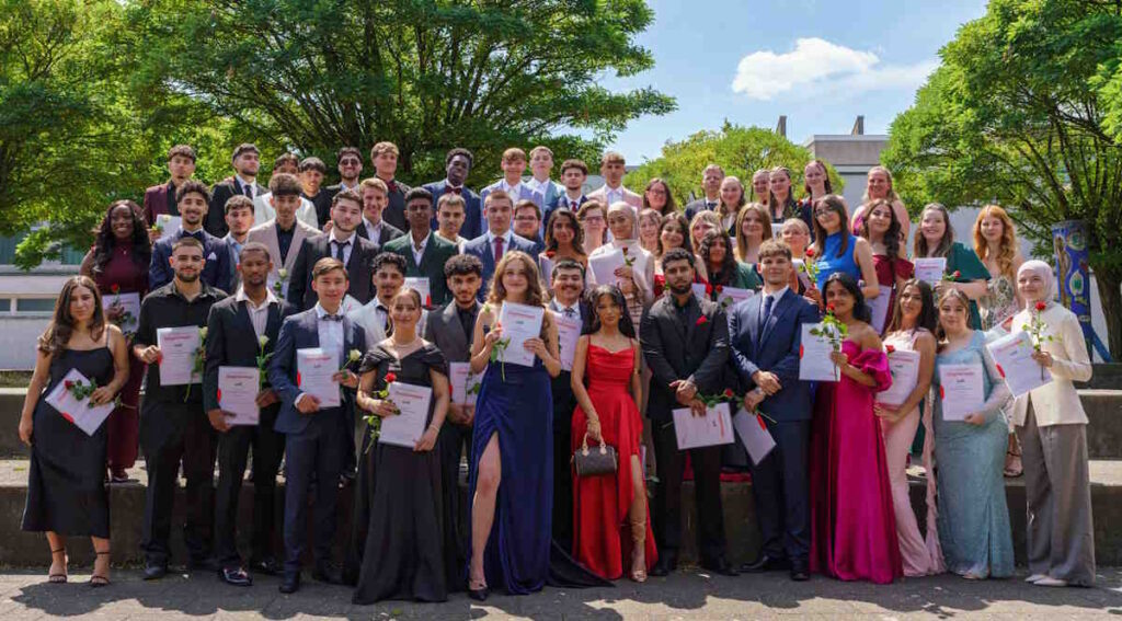Gruppenbild des Abiturjahrgangs 2025 der Gesamtschule Meiderich nach der Zeugnisverleihung. Die festlich gekleideten Schülerinnen und Schüler haben sich in 5 Reihen aufgestellt und halten alle ihr Abiturzeugnis und eine Rose in der Hand.