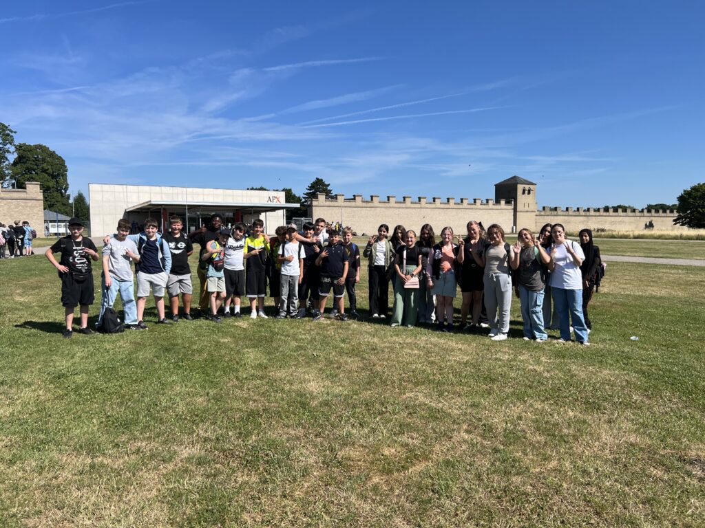 Eine größere Gruppe Jugendlicher steht auf einer Wiese vor dem Eingang des Archäologischen Parks Xanten (APX) und blickt in die Kamera. Im Hintergrund sind Reste römischer Mauern und Türme zu sehen.