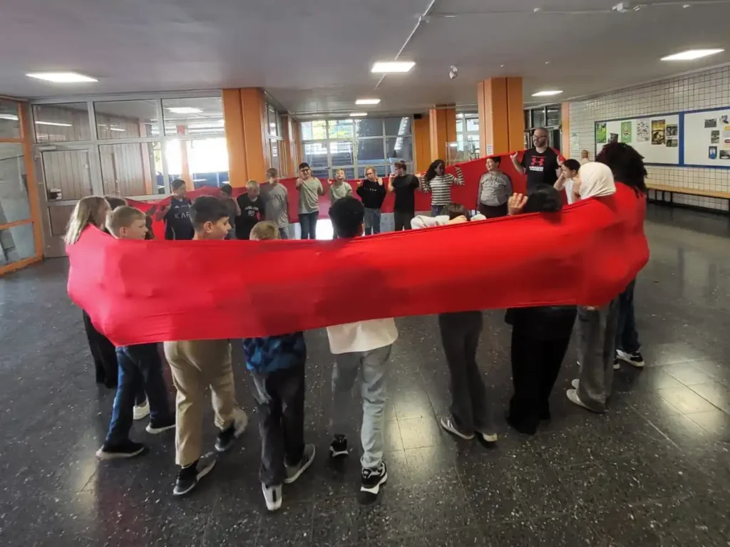 Eine Schülergruppe des Jahrgangs 5 bei einer Übung während des Sozialkompetenz-Trainings im Eingangsbereich der Schule. Die Schüler bilden einen geschlossenen Kreis und blicken sich an.