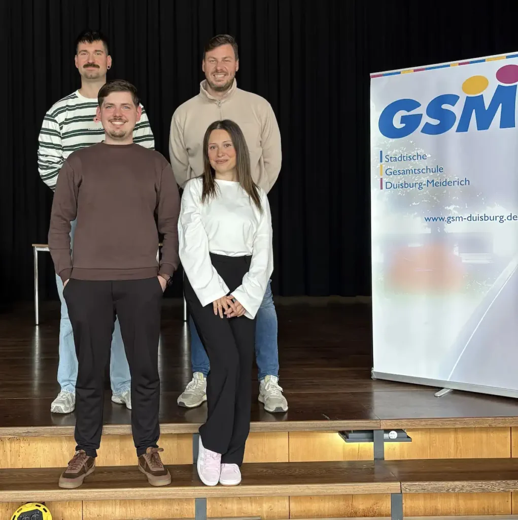 3 Lehrer und 1 Lehrerin stehen vor einem Plakat mit dem Logo der Gesamtschule Meiderich.